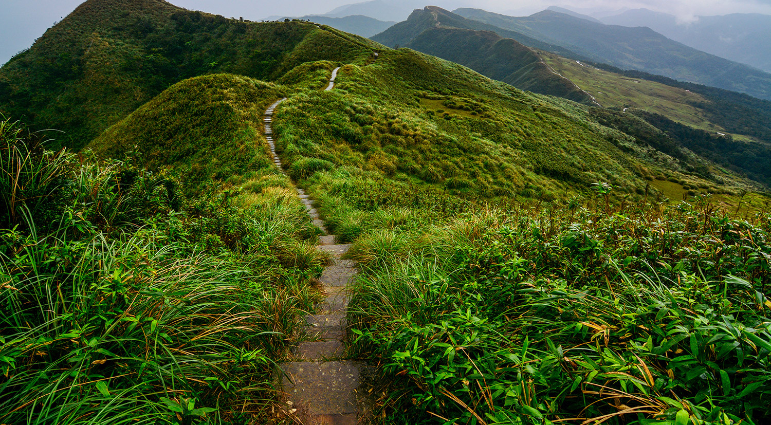 A picture of a big lush green mountain with other mountains in the background. There is a path of stairs leading down the mountain and far into the distance.
