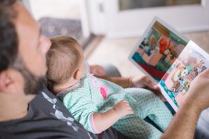 A father sitting with a baby on his lap. The father is reading a book to the baby. The father is wearing a black tshirt and the baby is in a teal and pink onesie.